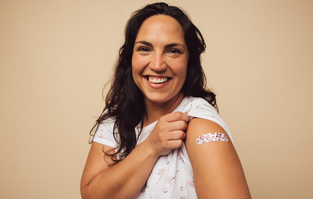 Portrait of a female smiling after getting a vaccine. Woman holding up her sleeve and showing her arm with bandage after receiving a flu shot.